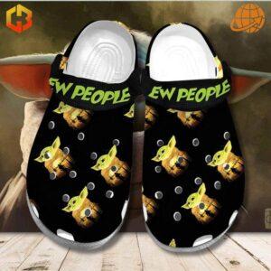 Ew People Baby Yoda Crocs Shoes displayed on a wooden surface with a blurred background, showcasing the black color, Baby Yoda illustrations, and green "EW PEOPLE" text on the strap.