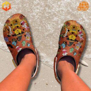 Feet wearing FRIENDS Winnie-the-Pooh Crocs Shoes on a sandy beach with ocean waves.