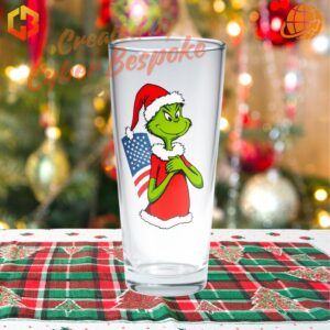 A festive beer glass featuring the Grinch holding a US flag, wearing a Santa hat, with 'Christmas 2024' text and patriotic design elements