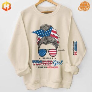 A beige sweatshirt featuring a bold graphic of a woman with a bun, wearing an American flag-patterned headband and sunglasses, accompanied by the text, “I’m Still A Trump Girl, I Make No Apologies,” and “Trump ’24” printed on the sleeve.