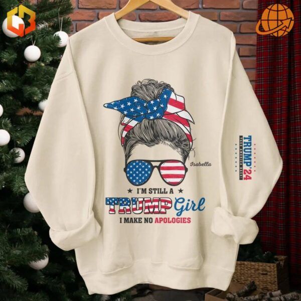 Beige sweatshirt featuring a detailed illustration of a bun hairstyle, sunglasses, and a headband with stars and stripes, accompanied by the slogan, 'I’m Still A Trump Girl, I Make No Apologies