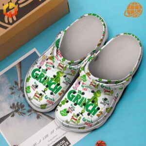 White The Grinch Clogs with festive illustrations, placed beside a photo of palm trees on a blue surface.